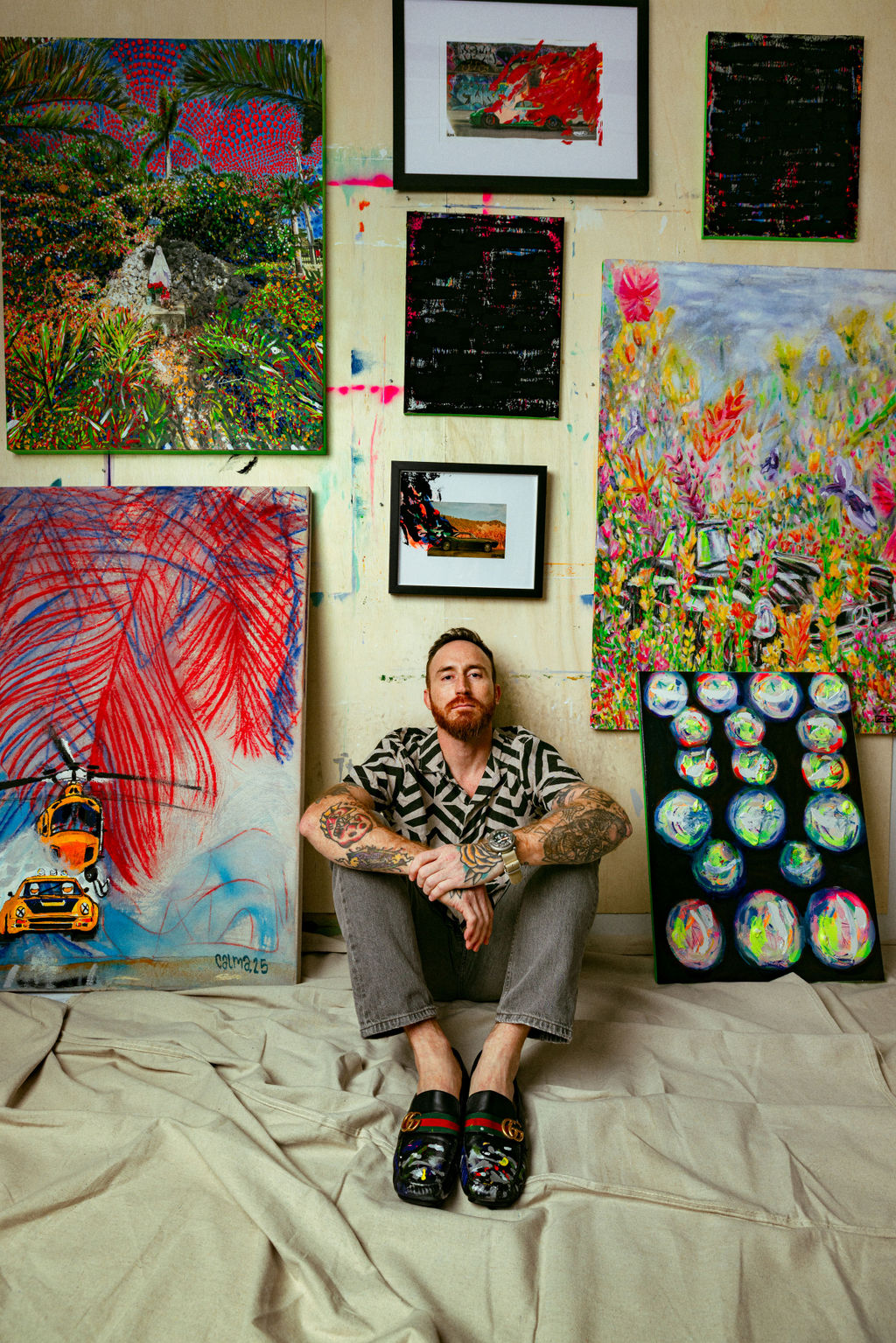 a red head bearded tattooed artist sits on the ground with paint dripped gucci slippers surround by his custom made art
